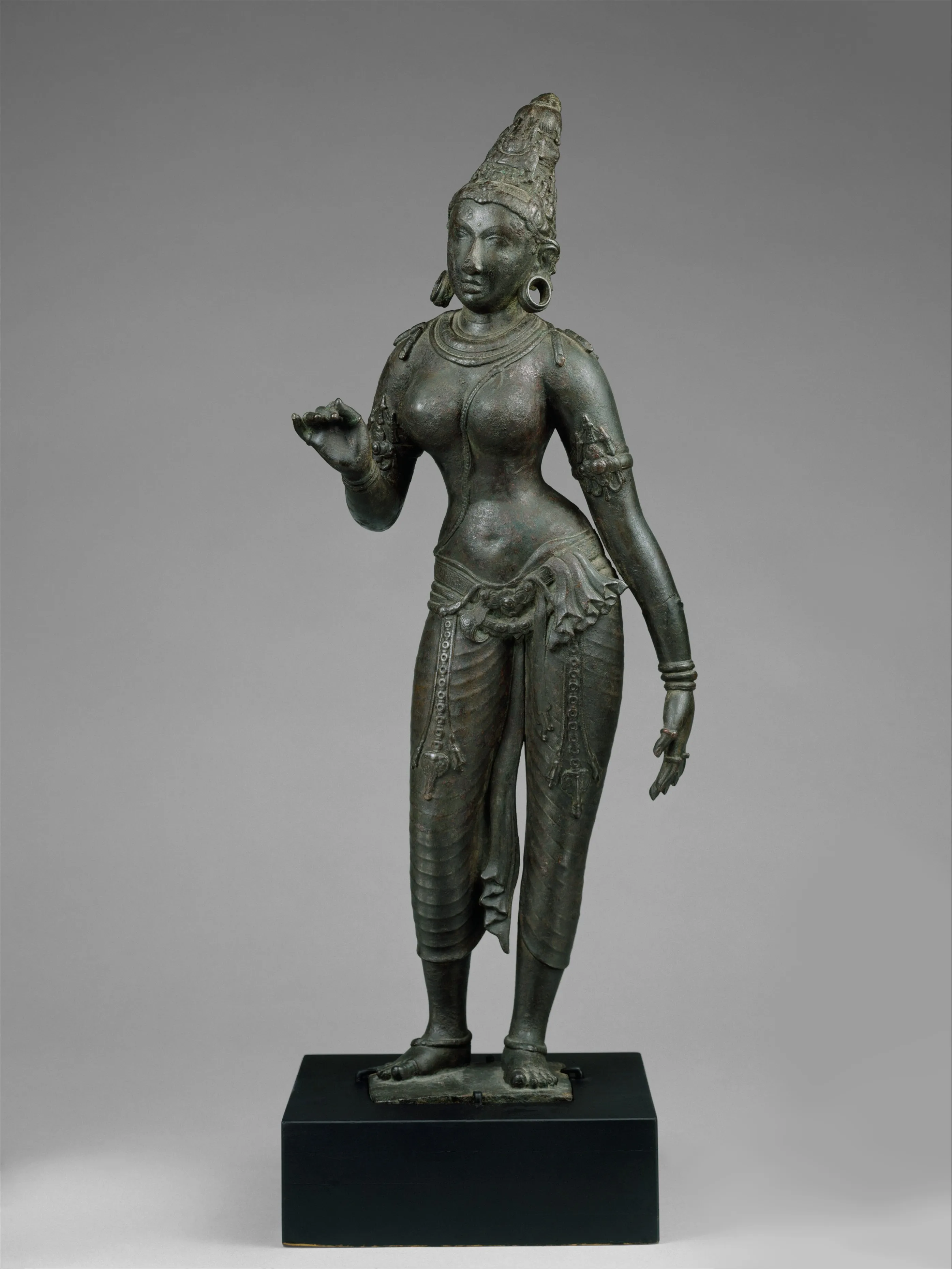சாம்பல் பின்னணியில் நிற்கும் சோழர் கால உலோக பெண் சிற்பம்